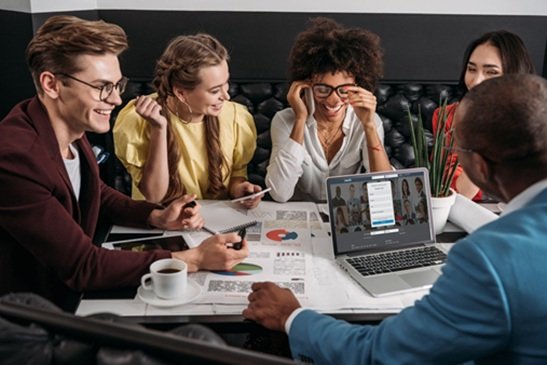 Equipe diversa reunida em café analisando gráficos de marketing com notebook em videoconferência durante uma reunião colaborativa.